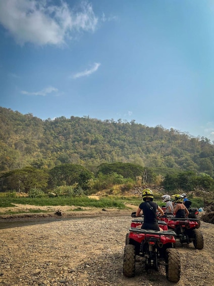 People riding red ATVs on a rocky path with a forested hill in the background. 3