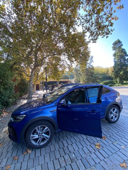 A blue car parked on a paved area with trees and a building in the background. 3