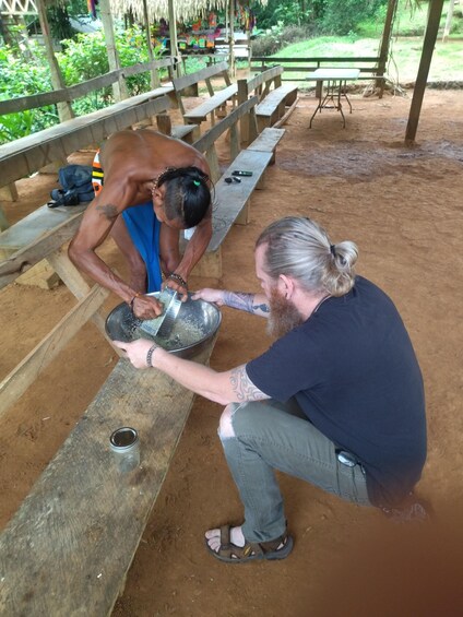 Two people at a wooden table outdoors, one pouring liquid from a metal container into a bowl. 1