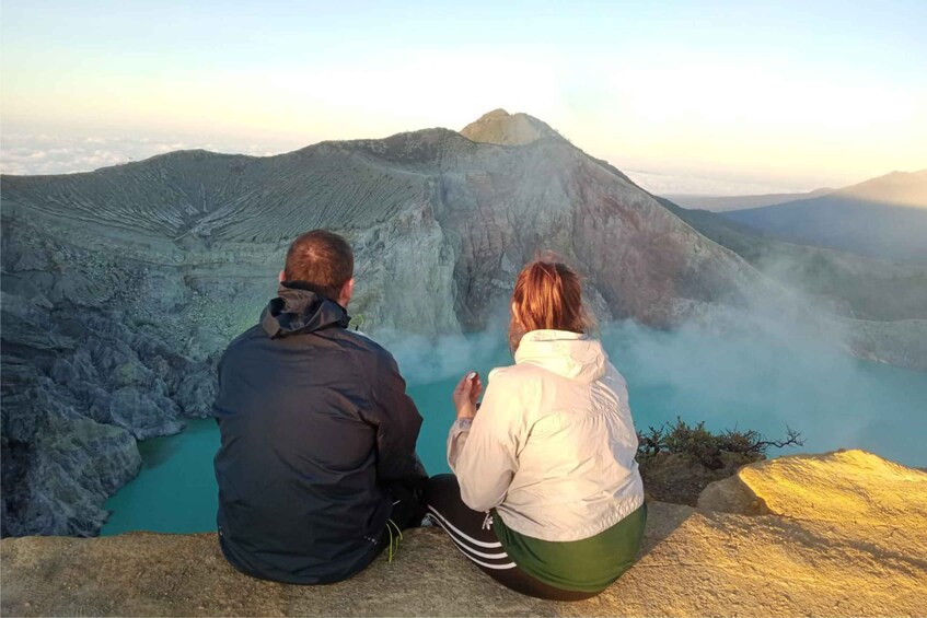 Two people sitting on a rock overlooking a volcanic crater with a blue lake. 2