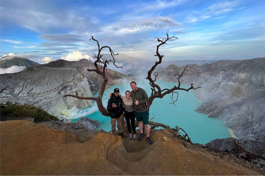 Three people standing on a rocky outcrop with a view of a turquoise lake and a barren tree in the foreground. 4