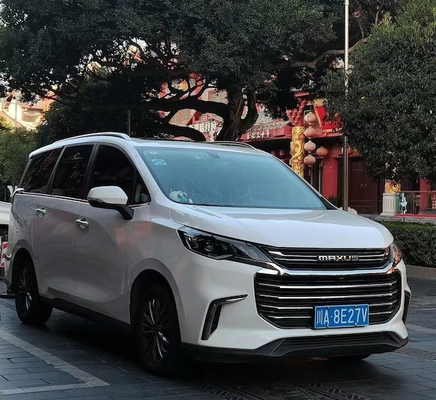 A white MAXUS SUV parked on a paved area with trees and a red building in the background. 4