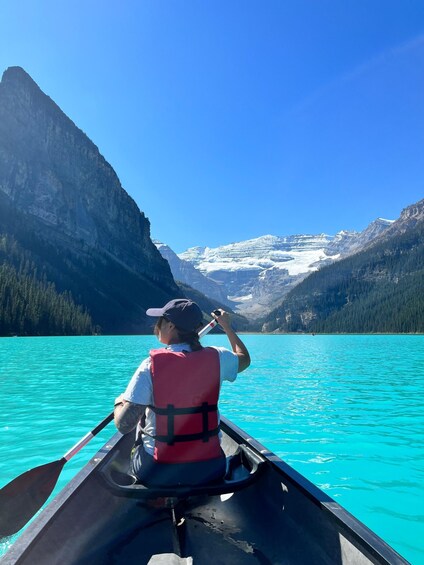A person in a canoe on a turquoise lake, wearing a life jacket and a cap, with mountains and snow-capped peaks in the background. 3
