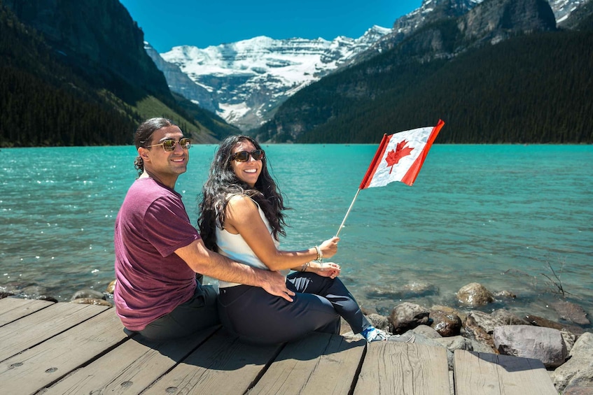 Two people sitting on a wooden dock by a turquoise lake with snow-capped mountains in the background. 1