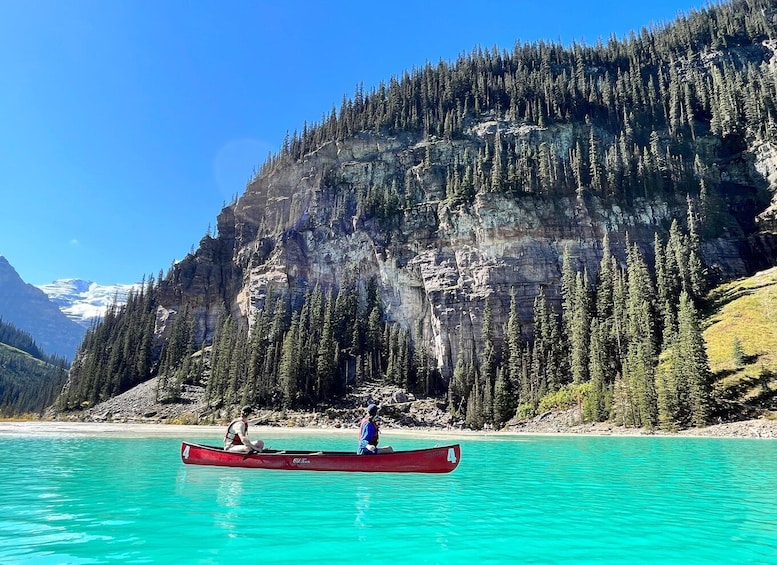 A red canoe on turquoise water with two people paddling, surrounded by a forested mountain and a clear sky. 4