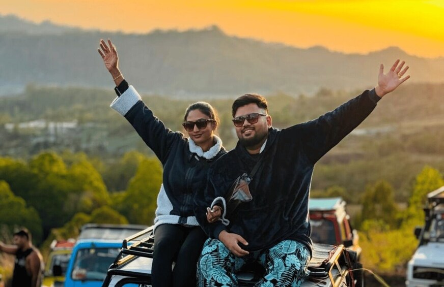 Two people on a motorcycle with their arms raised, a sunset in the background, and a hilly landscape. 4