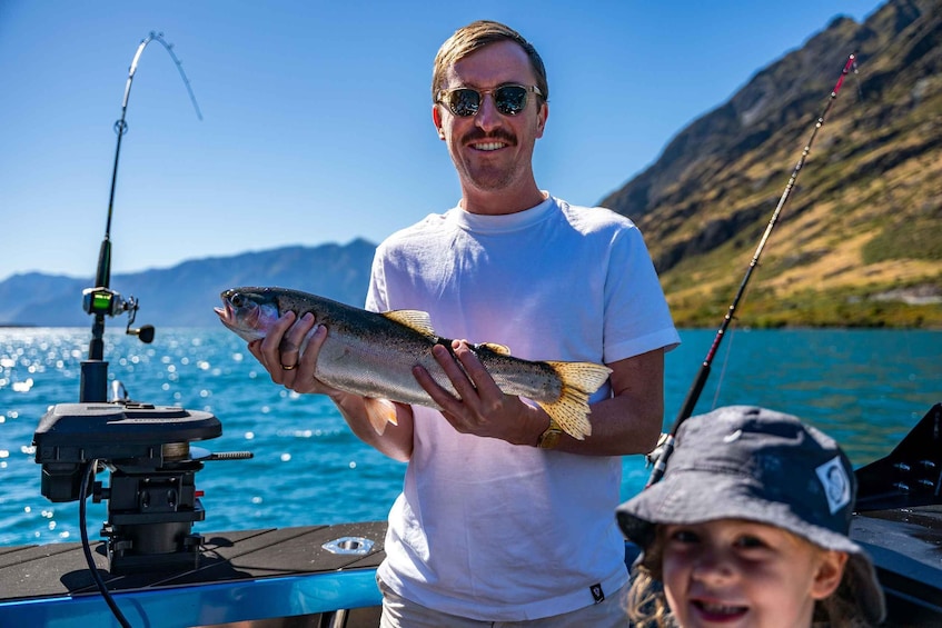 A person holding a fish on a boat with fishing rods and a mountainous landscape in the background. 1