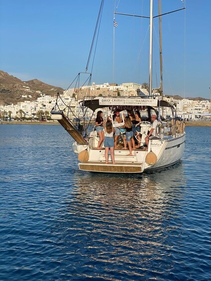 A sailboat named "Natalina" with passengers on board, docked near a coastal town with buildings in the background. 3