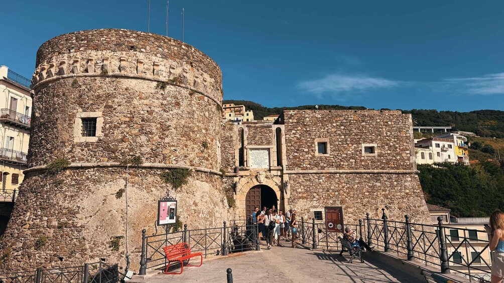 A historic stone castle with a round tower, a gate, and a paved walkway with benches. 3