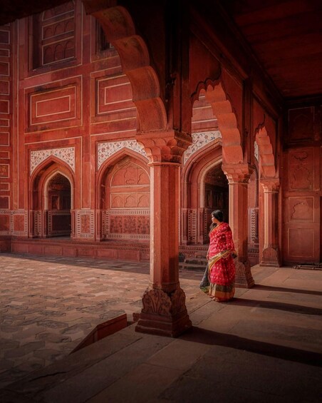 A woman in traditional attire walking through an ornate architectural space with red sandstone columns and detailed carvings. 3