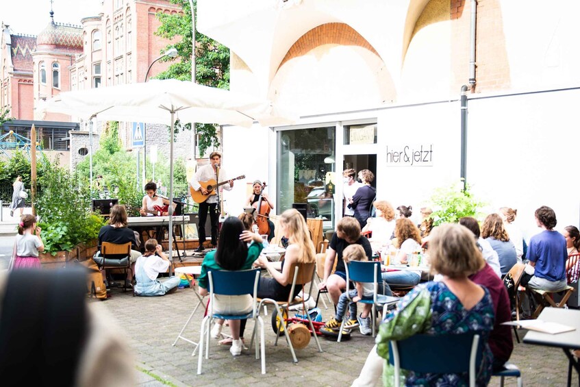 A group of people sitting outdoors, some playing musical instruments, with a building in the background. 2