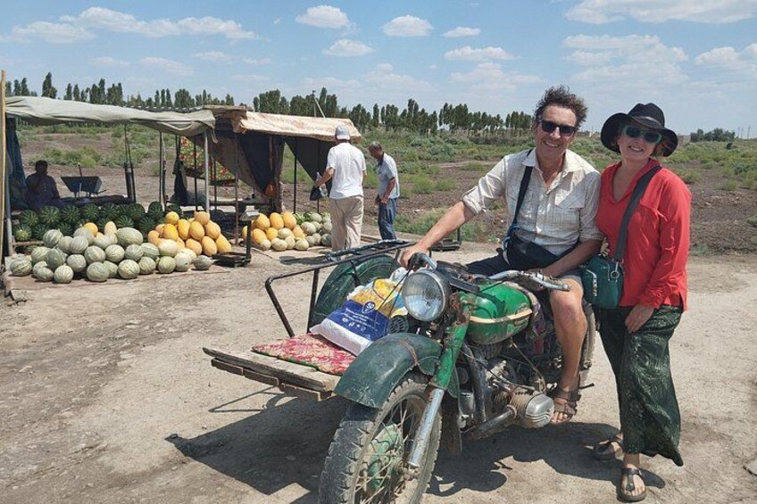 A man on a motorcycle with a sidecar, a woman standing beside him, and a market stall with melons in the background. 2