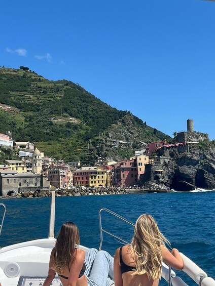 Un bote con dos personas a bordo, un pueblo costero con edificios coloridos y una colina rocosa con una torre. 1