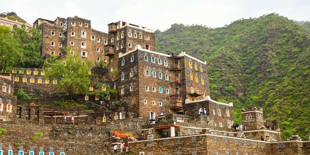 A multi-story stone building with balconies and windows, surrounded by lush greenery and hills. 1