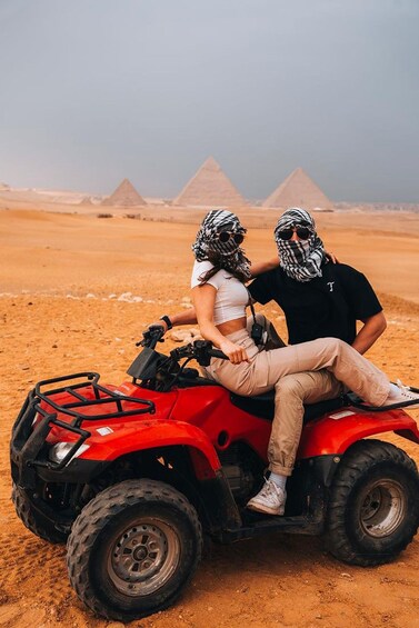 Two people on a red ATV in a desert with pyramids in the background. 1