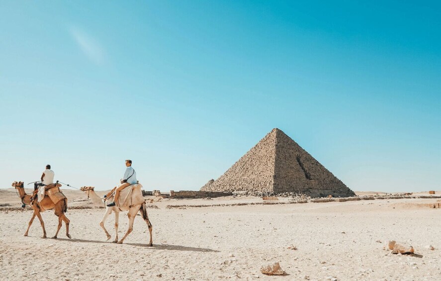 Two people riding camels in a desert with a large pyramid in the background. 2