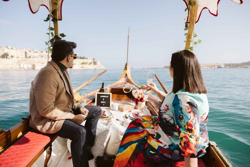 Two people on a boat with a table set for a meal, surrounded by water and a coastal town in the background. 2