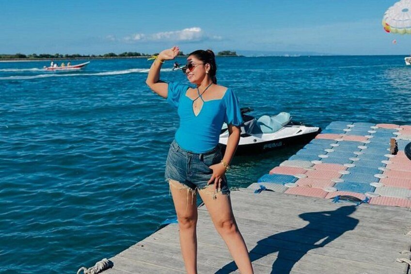 A person standing on a wooden dock by a body of water, with a boat and a parasail in the background. 2