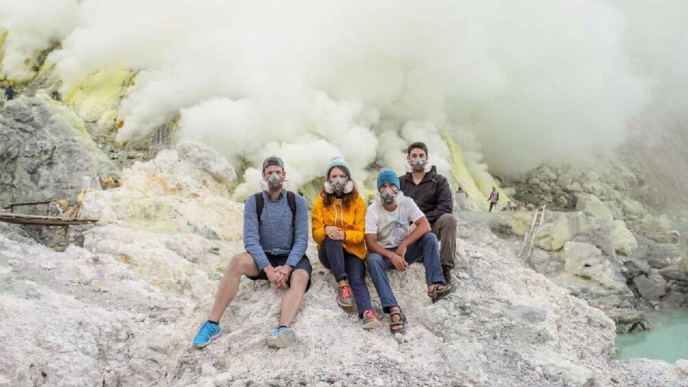 Four people sitting on a rocky outcrop with a geothermal vent emitting steam in the background. 3