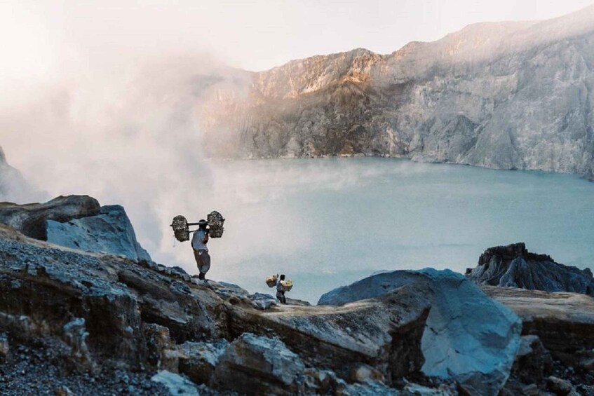 A person carrying a large load across a rocky terrain near a lake with mountains in the background. 1