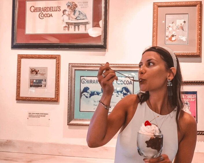 A person holding a glass with a dessert, standing in front of framed advertisements for Ghirardelli's Cocoa. 3