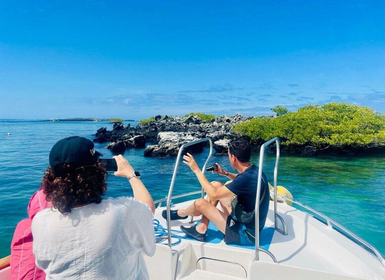 Two people on a boat taking photos of a rocky shoreline with clear blue water. 2