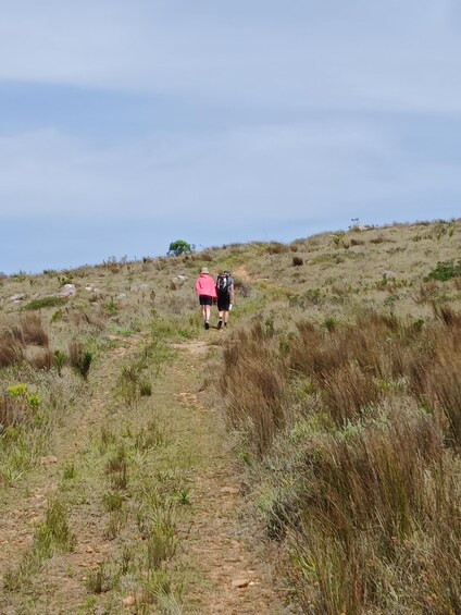Two people hiking on a dirt path through a grassy landscape with sparse vegetation and a clear sky. 3