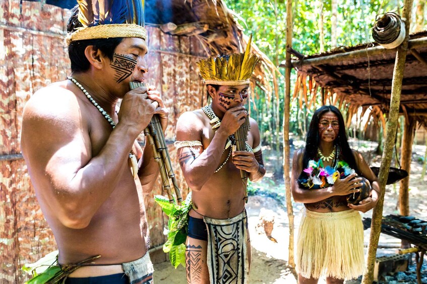 Two men with tribal tattoos and headdresses, one holding a plant, standing in a rustic outdoor setting with a thatched-roof structure in the background. 1