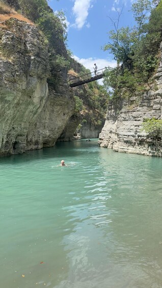 A person walking on a bridge over a river, surrounded by rocky cliffs and greenery. 3