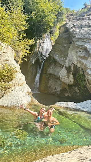 Two people in a clear pool with a waterfall in the background. 1