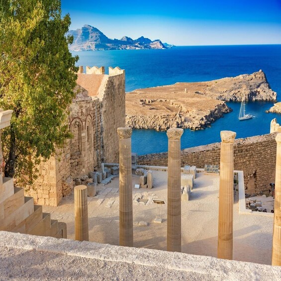 Ancient ruins with columns, a tree, and a coastal view with a sailboat. 3