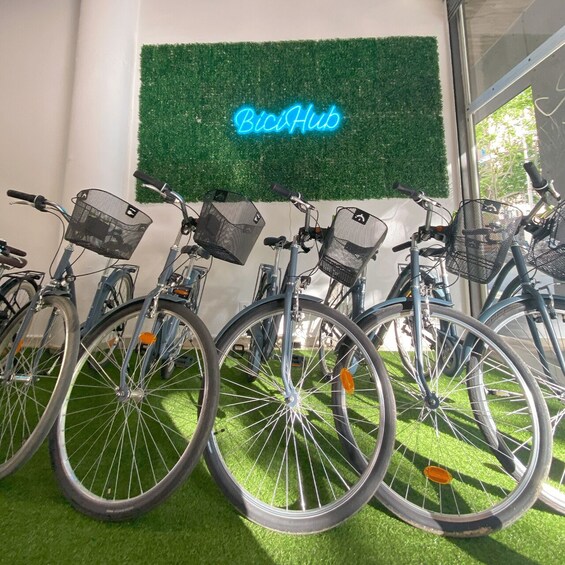 A row of bicycles with baskets, a neon sign reading 'BiciHub,' and a green artificial grass floor. 2