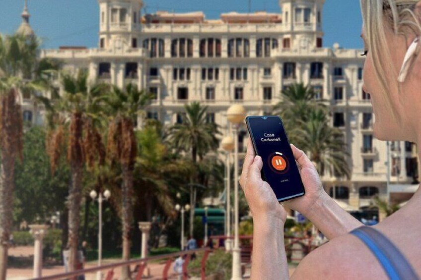 Une personne tenant un smartphone affichant l’interface de l’application « Casio Carbonell », avec des palmiers et un grand bâtiment à l’architecture classique en arrière-plan. 5