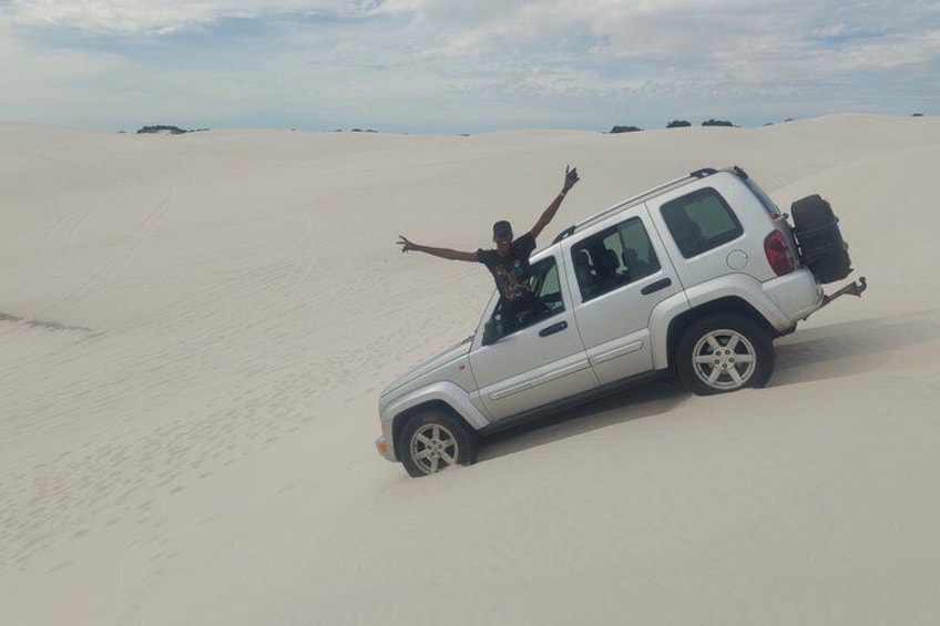 En vit SUV står på en sanddyn, och en person har armarna lyfta mot skyn. 4