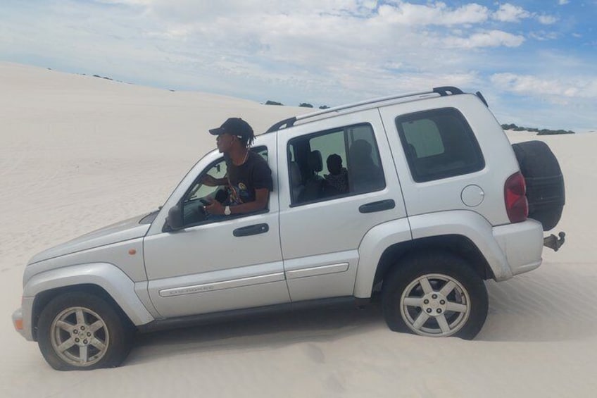 En vit SUV står på en sandstrand och någon kör den. 3