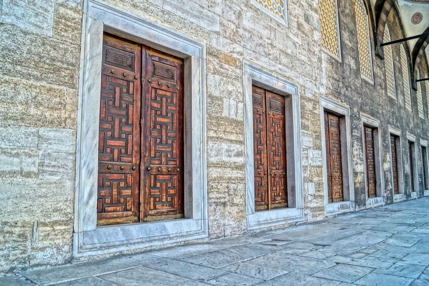 A row of ornate wooden doors with geometric patterns, set in a stone building with arched windows. 5