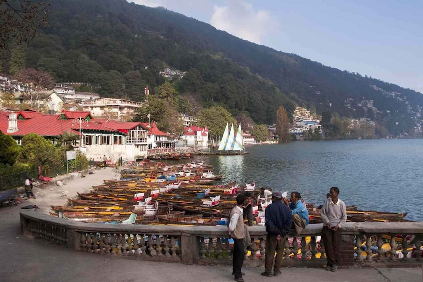 A lakeside scene with boats docked, people by the water, and buildings on the hillside. 1