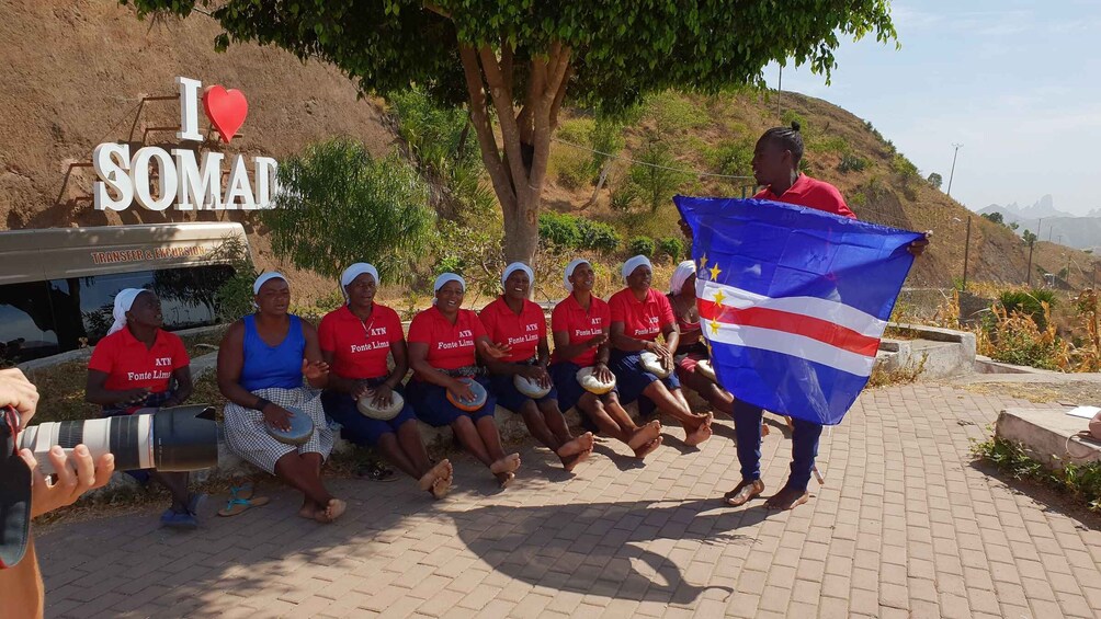 A group of people sitting on a bench under a tree, holding drums, with a sign that says "I love SOMAL". 5