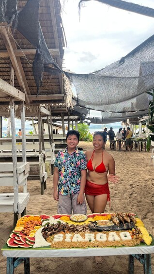 A beachside dining setup with a variety of food items, including seafood and fruits, under a thatched roof. 1