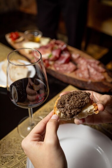 A person holding a piece of bread with a spread, a glass of red wine, and a charcuterie board in the background. 4