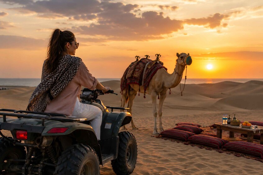 Una persona monta un cuatrimoto en el desierto al atardecer, con un camello cerca. 2