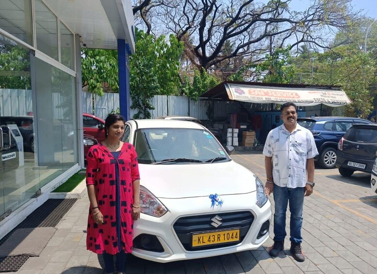A white Suzuki car with a blue ribbon on the hood, parked in front of a dealership with a signboard in the background. 3