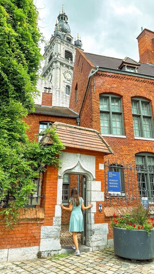 A woman in a light blue dress standing at the entrance of a red brick building with a white door and a clock on the wall. 3