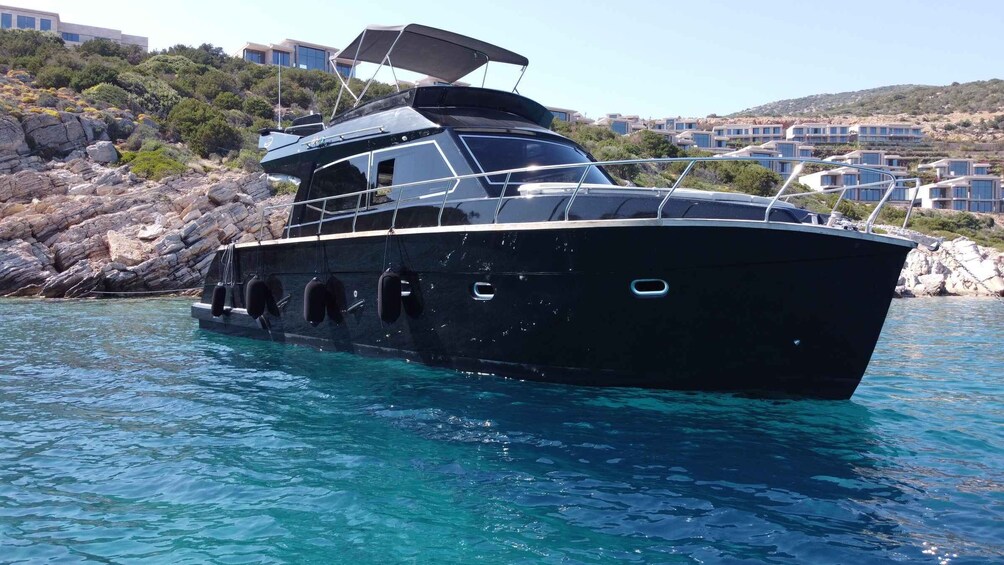 A black boat with a covered upper deck, anchored near rocky shores with buildings in the background. 1
