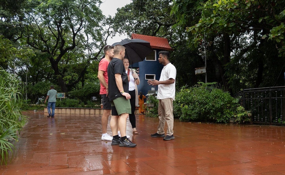 A group of people standing on a wet pavement in a park with trees and a small building in the background. 2