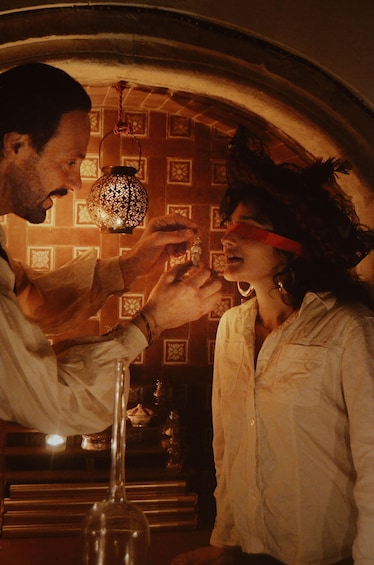 A man and woman in a dimly lit room with patterned wallpaper, a hanging lantern, and a wine glass on a table. 3