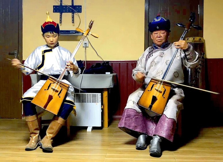 Two individuals playing bowed string instruments in a room with wooden floors and a white air conditioning unit. 3