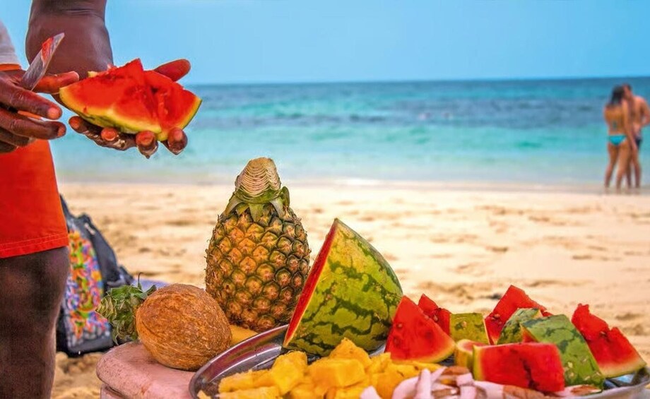 A beach with a fruit stand, including watermelon, pineapple, and coconut. 2