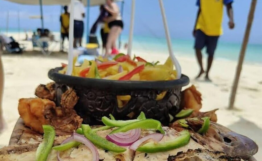 Grilled fish on a wooden board with sliced onions and peppers, a black pot with food, and a beach with people in the background. 3