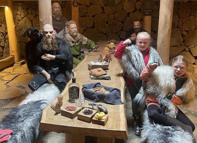 A group of people sitting around a wooden table with various food items and a bottle. 1